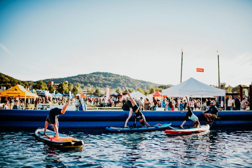 Three people on stand up paddleboards doing yoga. There is a festival happening in the background.