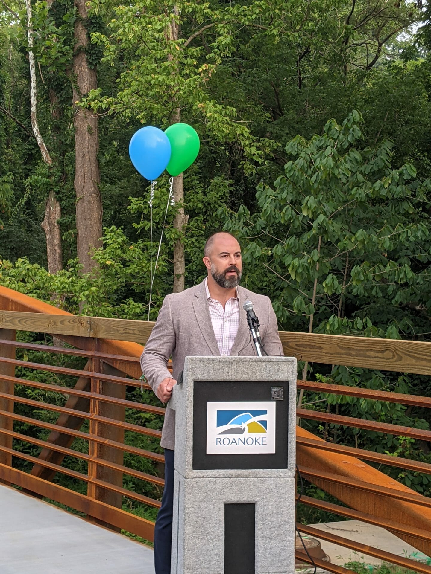 White man speaking at a podium outside with blue and green balloons in the background 