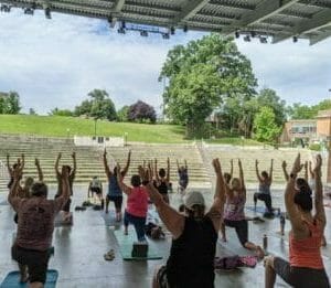 Several people on an outdoor stage practicing yoga with a green hill in the background