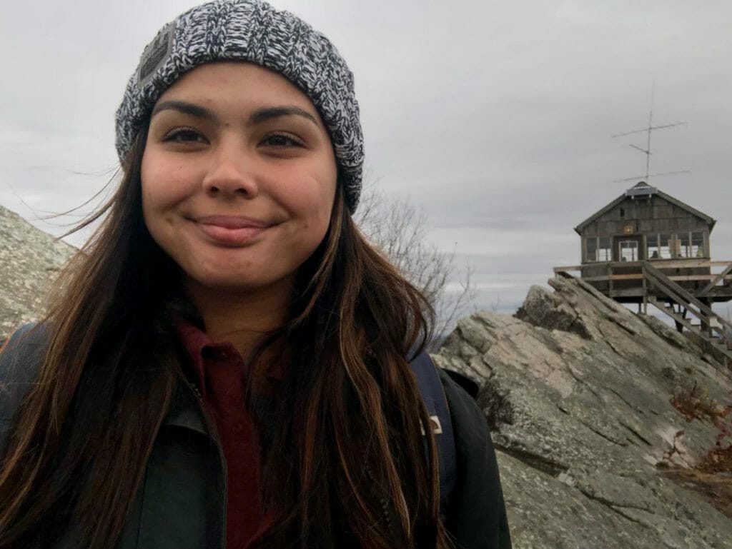 Young woman outdoors wearing a jacket and a beanie with a watch tower in the background