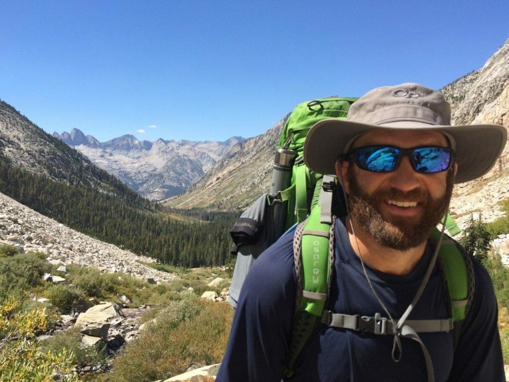 Man with beard, sunglasses, and bucket hat smiling at the camera with large mountain range in the background