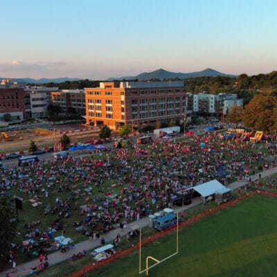 Aerial view of large crowd in a park with food trucks, stage, and bounce houses with blue ridge mountains in the distance