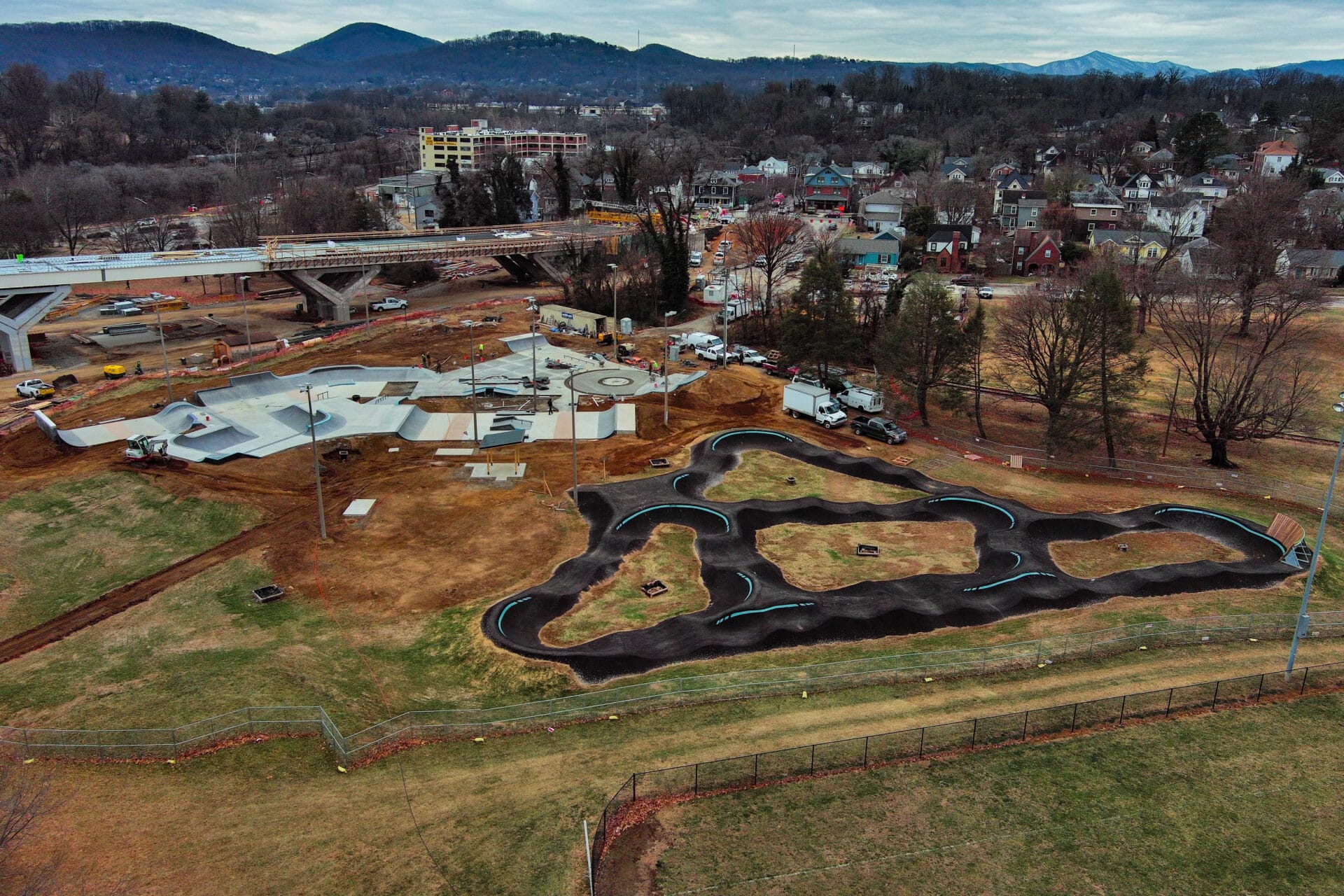 Aerial view of a large skatepark and pump track with bridge construction and mountains in background.