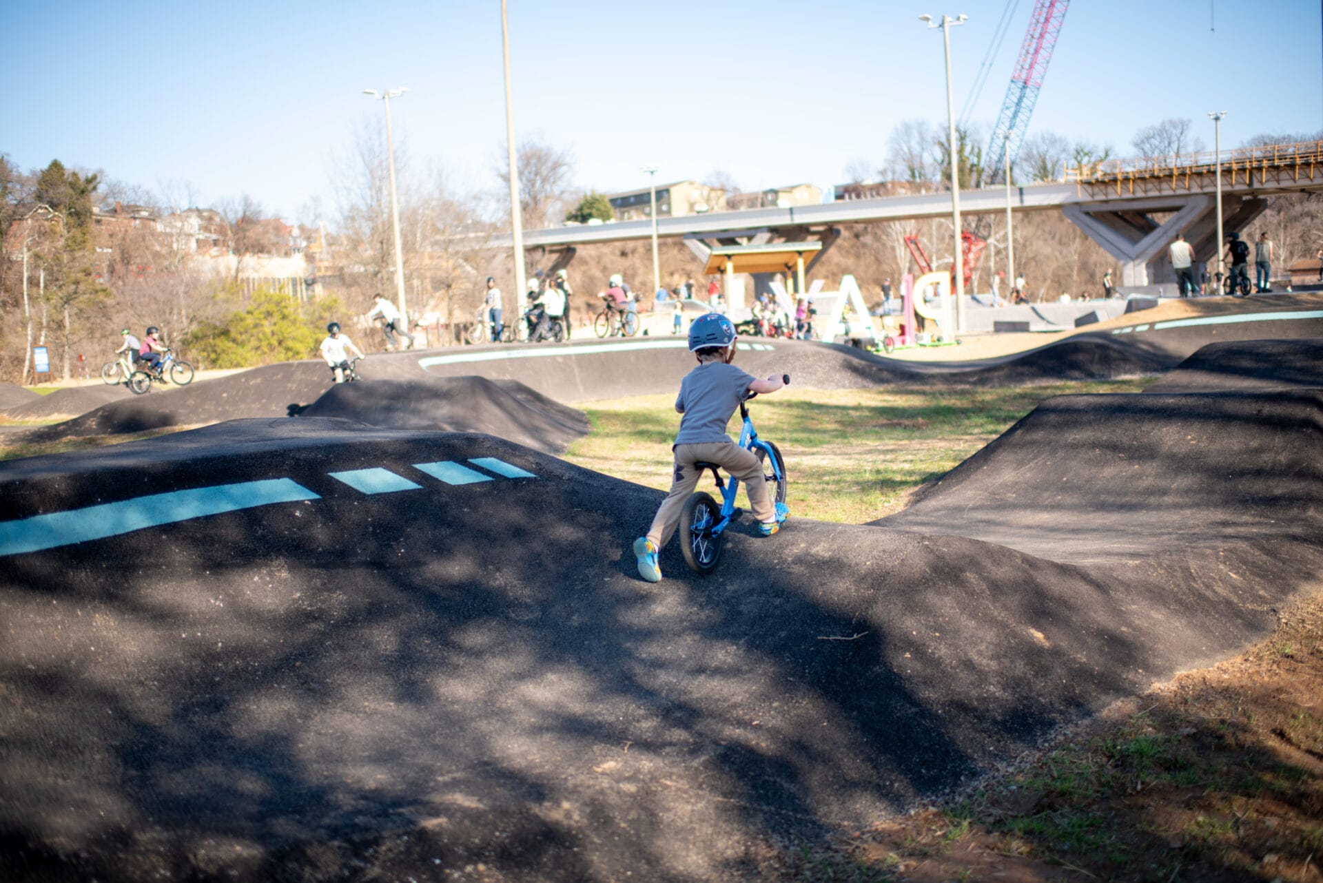 A child riders a balance bike up the incline of a large asphalt pump track.