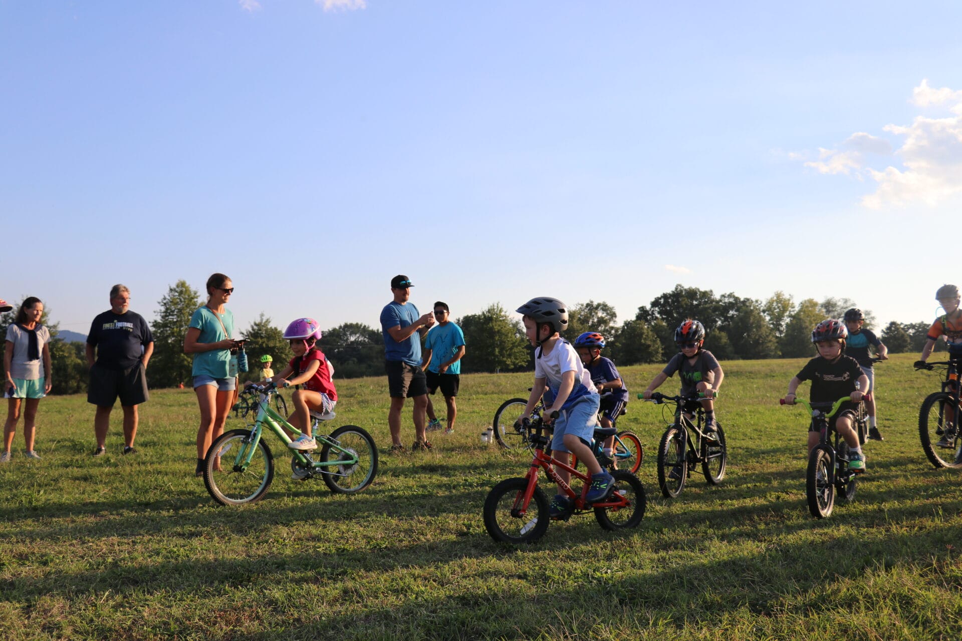 A group of children rides bikes in a park field as part of a youth bike race, with their parents supporting on the sidelines.