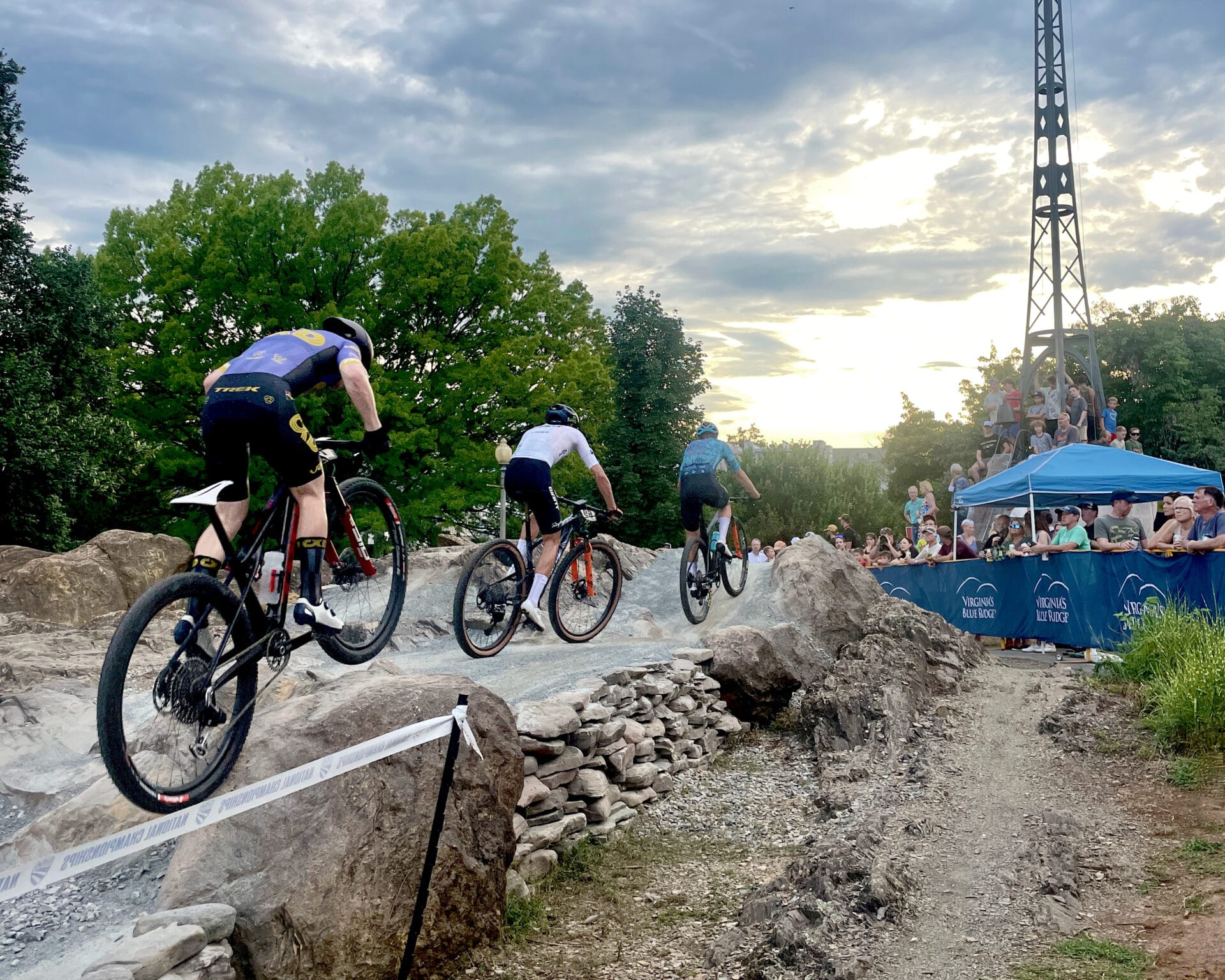 Cyclists ride over a rock feature in front of a large crowd at a USA Cycling race.