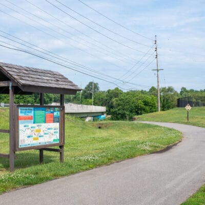 Kiosk and boat rack along a paved greenway near a river.
