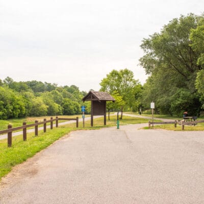Parking lot adjoining a paved greenway, with kiosk, trash can, and picnic table.