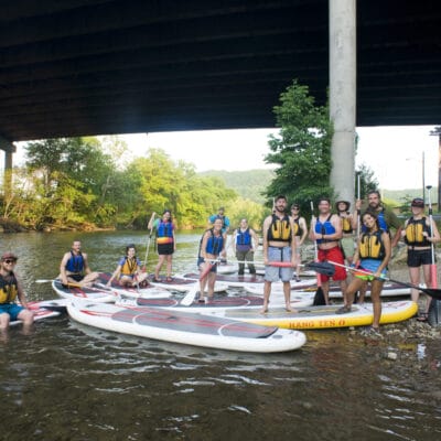 A group of paddleboarders wearing pfd's gathered along the shoreline of a river, posing for a photo.
