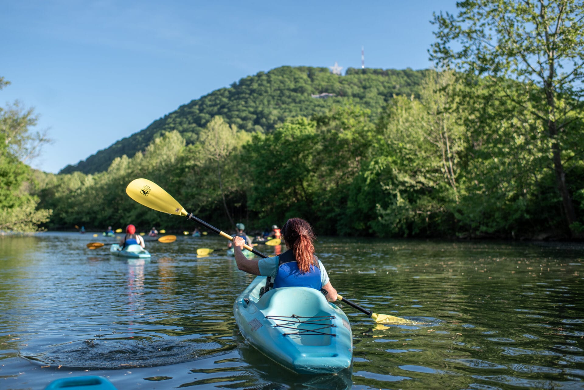 A woman paddles a kayak on a river with Mill Mountain and the Roanoke Star in the distance.