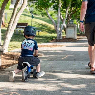 TinyTotTri26 850web A toddler on a balance bike rides beside their parent while competing in a 'triathlon' event for toddlers.