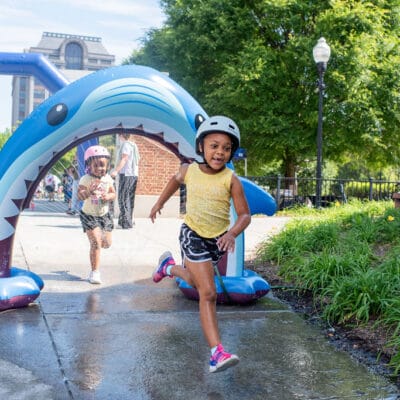TinyTotTri26 904web Two girls run through a sprinkler as part of a 'triathlon' event for toddlers and small children in a city park.